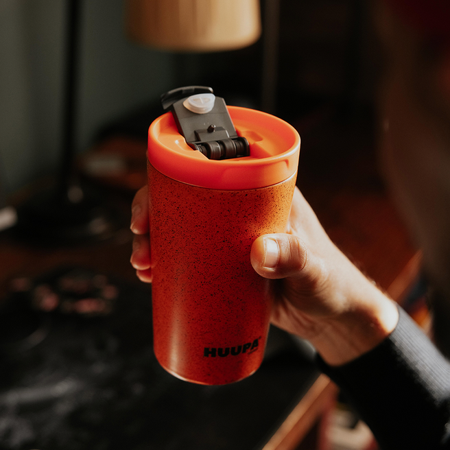 Mano sosteniendo termo naranja "Huupa" con fondo en casa borroso.