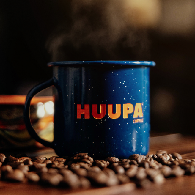 Taza azul peltre "Huupa" sobre granos de café mesa de madera y fondo borroso en casa.