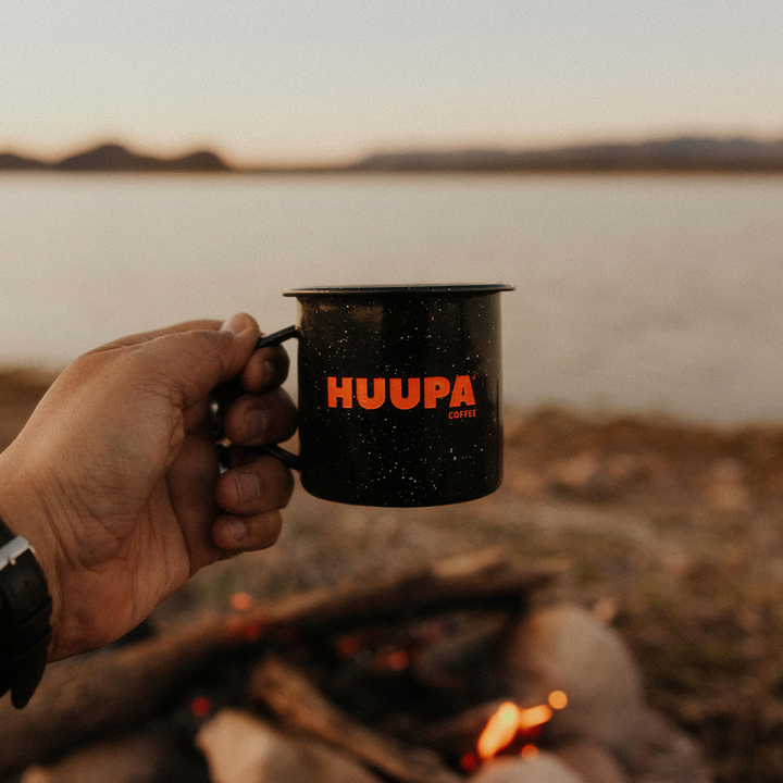 Mano sosteniendo taza negra peltre "Huupa" al exterior con vista al mar y fogata prendida.