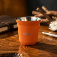 vaso medidor de espresso 80ml naranja marca HUUPA en mesa de madera y pluma en el fondo