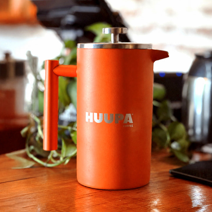 Prensa francesa naranja 1 L marca Huupa arriba de emsa de madera y plantas verdes en el fondo